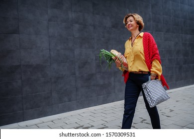 Charming caucasian fashionable senior woman carrying bag and groceries while passing by gray wall.