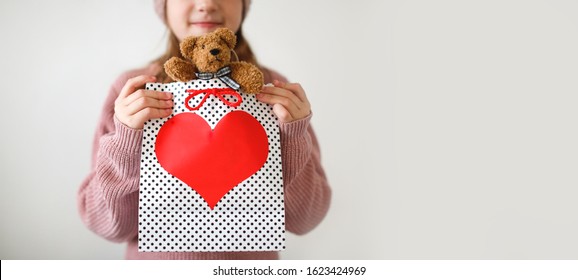 Charming Caucasian Kid Girl With A Soft Teddy Bear In A Gift Bag With A Big Heart, Concept For Love, Gift And Valentine's Day. Girl In A Sweater And Knitted Hat