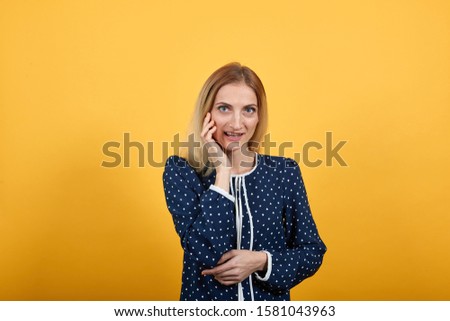 Charming caucasian young lady looking at camera, keeping hand on cheek over isolated orange background in blue shirt with white polka dot. Lifestyle concept