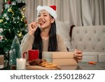 A charming, cheerful Asian woman enjoys eating cookies at a coffee table in the living room on the Christmas holiday, celebrating the holiday alone at home.