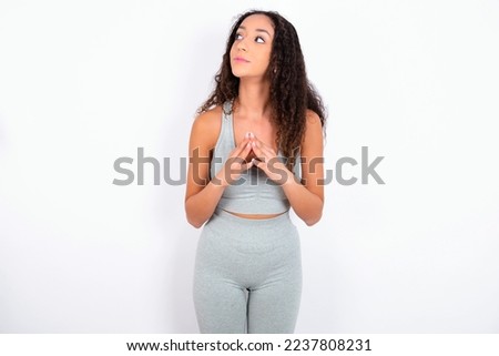 Charming cheerful Beautiful teen girl with curly hair wearing grey sport set over white background making up plan in mind holding hands together, setting up an idea.