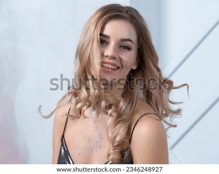 Charming cheerful blonde with long wavy hair and sparkling makeup. Beautiful young woman with sequins on her face