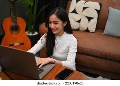 Charming And Cheerful Young Asian Female Freelancer Or College Student Typing On Notebook Keyboard, Working On Her Tasks On Laptop Computer At Home.