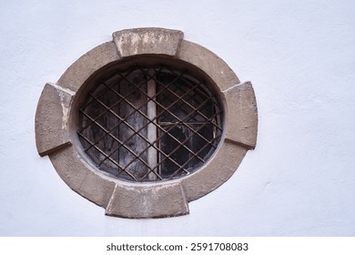 Charming circular window featuring rusted metal grates, set in an aged concrete wall, evokes historical architectural perspective and timeless detail. The white wall creates a contrasting background.