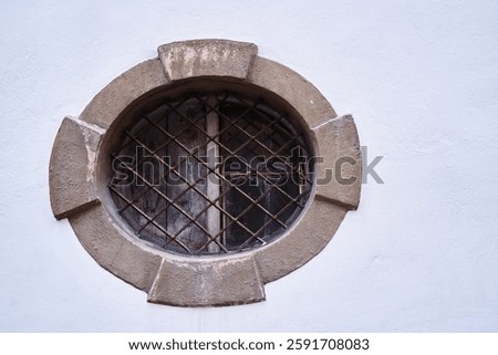 Charming circular window featuring rusted metal grates, set in an aged concrete wall, evokes historical architectural perspective and timeless detail. The white wall creates a contrasting background.