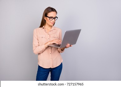 Charming Clever Lady In A Beige Formal Smart Casual Shirt Is Standing On The Pure Grey Background With Laptop In Her Hands, Smiling, Looking At It While Browsing, Typing, Writing, Wearing Black Specs