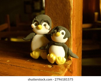 A charming, close-up shot of two small penguin figurines handcrafted from modeling clay or playdough. The cute figures, featuring classic black, white, and yellow details, are sitting together on a warm, rustic wooden surface under soft indoor lighting, creating a cozy and creative atmosphere.