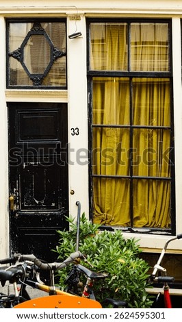A charming close-up of a traditional Dutch house facade with a worn black door, large window with yellow curtains, and several bicycles parked in front.