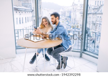 Charming couple laughing while sitting in a cafe with their modern phone. The girl waves at the camera, chatting, video conference online, using free Wi-Fi.