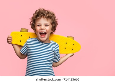Charming curly boy holding yellow longboard and looking away on pink background. 