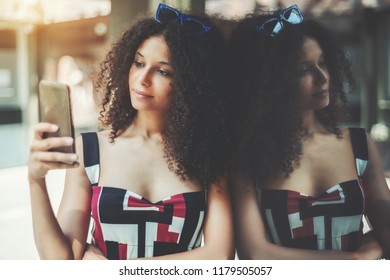 A charming curly caucasian girl is typing a message on her smartphone while leaning against glass wall; dazzling young Moroccan woman is using her cell phone while waiting for her friend outdoors - Powered by Shutterstock