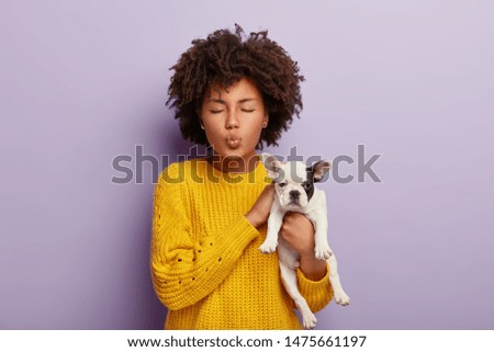 Charming curly haired woman takes care of adorable pet, enjoys pleasure from playing with favourite dog, keeps eyes shut, spends weekend with cute puppy stands over purple background. Domestic animals