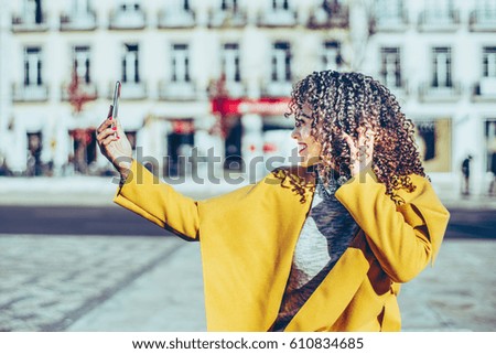 Charming curly smiling Brazilian woman in yellow coat and sweater making selfie on her smart phone while standing on sunny street with blurred city road and facade of residential house in background