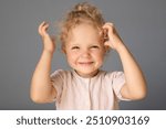 Charming cute cheerful toddler girl with blonde wavy hair wearing rose sleeper sitting on bed on white sheet looking at camera with toothy smile and happy face isolated over gray background