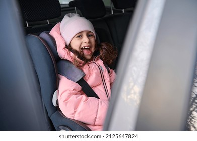 Charming Cute Little Child, Cheerful Girl In Pink Warm Clothes Smiles Sitting Fastened With A Secure Seat Belt In A Modern Car During A Family Winter Trip By Car. Concept Of Safe Travel With Children