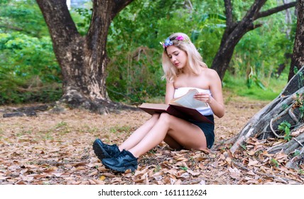 Charming Cute Young Woman Sitting On Fallen Autumn Leaves In The Park While Reading A Book In Her Hands. Cheerful Teen Girl Blonde Hair With Flower Learning In The Book Relaxed Under Tree.