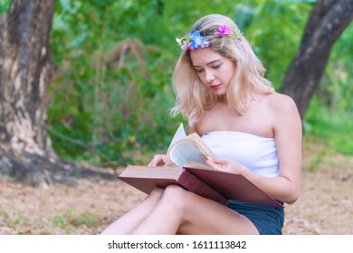 Charming Cute Young Woman Sitting On Fallen Autumn Leaves In The Park While Reading A Book In Her Hands. Cheerful Teen Girl Blonde Hair With Flower Learning In The Book Relaxed Under Tree.