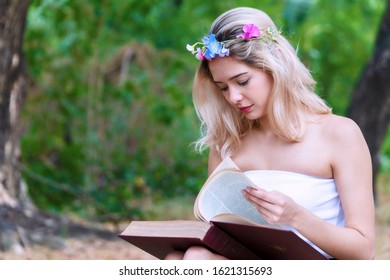 Charming Cute Young Woman Sitting On Fallen Autumn Leaves In The Park While Reading A Book In Her Hands. Cheerful Teen Girl Blonde Hair With Flower Learning In The Book Relaxed Under Tree.