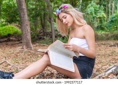 Charming Cute Young Woman Sitting On Fallen Autumn Leaves In The Park While Reading A Book In Her Hands. Cheerful Teen Girl Blonde Hair With Flower Learning In The Book Relaxed Under Tree.