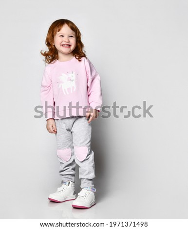 Charming cutie toddler smiling red-haired little girl in sportswear . Female child in pink raglan with cute unicorn print design and sports pants. Headshot portrait