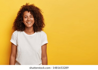 Charming dark skinned teenage girl with crisp Afro hair, expresses cheer and good mood, smiles happily, shows perfect teeth, wears white everyday t shirt, models against yellow studio background