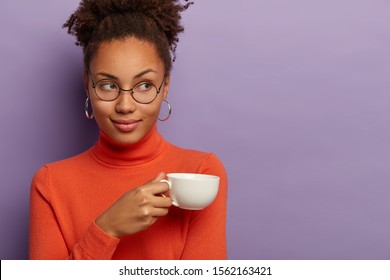 Charming dark skinned woman with curly crisp hair, drinks coffee or tea, holds white mug, wears spectacles and orange turtleneck, looks aside, isolated over purple background, free space right