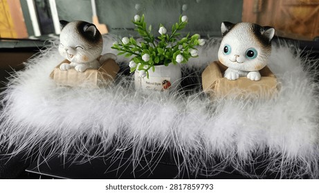 A charming decorative display featuring two adorable kitten figurines placed beside a small potted plant with delicate white blossoms, all surrounded by soft white feathers. The smiling cat ornaments, expressive blue eyes, and cozy arrangement create a warm, playful, and inviting atmosphere perfect for home décor, nursery themes, gift ideas, or seasonal decorations. This composition blends cuteness with minimalist aesthetics, making it an excellent choice for lifestyle, interior design, decorative accessories, and creative background concepts. The soft textures and balanced composition evoke comfort, happiness, and a touch of whimsy.
