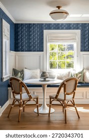 A charming dining nook featuring blue patterned wallpaper, white wainscoting, and a round white table accompanied by wicker chairs with white cushions. The space is illuminated by natural light from a window with Roman shades and a ceiling light fixture.