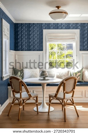 A charming dining nook featuring blue patterned wallpaper, white wainscoting, and a round white table accompanied by wicker chairs with white cushions. The space is illuminated by natural light from a window with Roman shades and a ceiling light fixture.