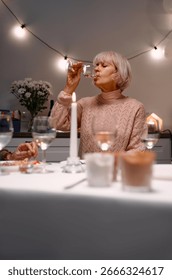 A charming dinner table moment featuring a warmly lit ambiance with senior woman appreciating a glass of white wine. The tabletop is accented with lit candles, glassware, and cozy decor.