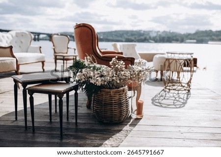 Charming dockside wedding lounge setup with vintage armchairs, small tables, and a basket of white wildflowers overlooking the water on a sunny day.