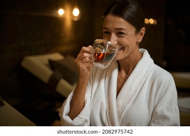 Charming elegant middle aged European woman in white bathrobe, drinking hot healthy herbal tea drink, sitting on chaise lounge by jacuzzi, enjoying spa procedures in spa salon - Powered by Shutterstock