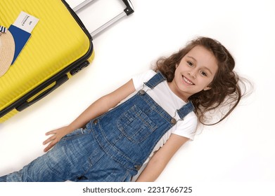 Charming European little child, adorable baby girl passenger wearing blue denim overalls, lies on white background near her boarding pass on yellow suitcase, smiles a beautiful toothy smile to camera