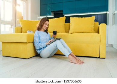 Charming Exuberant Woman Holding Her Ipad In Hands ,sitting On The Floor Near Light Green Couch And Enjoying Coffee In The Cup In The Contemporary Cuisine