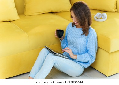 Charming exuberant woman holding her ipad in hands ,sitting on the floor near light green couch and enjoying coffee in the cup in the contemporary cuisine