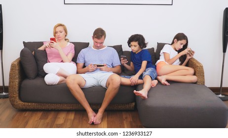 Charming Family, Mom, Dad, Daughter And Son Are Watching TV In The Living Room Together, Everyone Is Looking Into His Phone