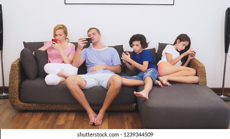 Charming Family, Mom, Dad, Daughter And Son Are Watching TV In The Living Room Together, Everyone Is Looking Into His Phone