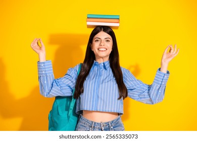 Charming female student in blue striped shirt balancing books on head against yellow backdrop - Powered by Shutterstock