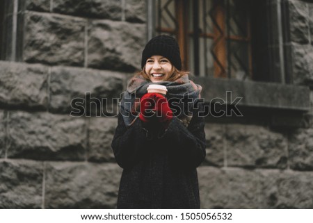 Charming female wearing warm coat, black hat, red gloves and scarf laughing and warming hands while holding cup of hot beverage, standing on background of gray building 