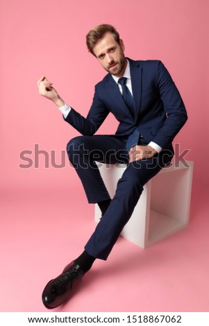 charming formal business man in navy suit is sitting and resting his hands on his laps with two fingers attached at one hand and looking at camera confident on pink studio background