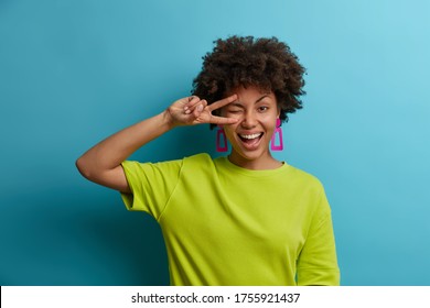Charming friendly African American woman makes peace sign over eye, smiles broadly and feels positive emotions, sends good vibes, wears bright green t shirt. People, emotions, summertime concept