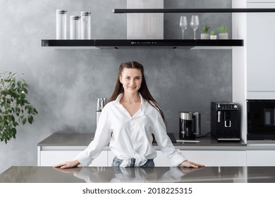 Charming friendly young woman standing at kitchen table in her new modern apartment and smiling at camera, happy female being ready to cook tasty food or meet guests in house - Powered by Shutterstock