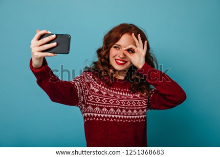 Charming ginger lady making selfie on blue background in christmas. Cheerful curly girl in new year sweater taking picture of herself.