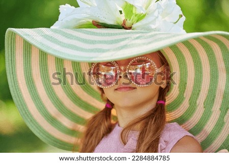 A charming girl child with wavy blonde hair wearing fashionable summer pink dress, stylish sunglasses and wide-brimmed hat walks on a street. Kid's summer fashion. Summer holidays.