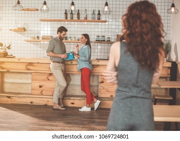 Charming Girl Is Flirting With Man And Smiling. She Is Touching Gift Present In Male Hands With Curiosity. Young Woman Standing In Front Of Them While Feeling Jealousy. Adultery Concept