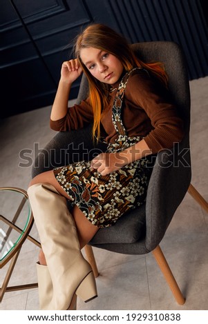 Charming girl with hair in posing in her mother's suede boots. Childhood, young actress. Full length shot, isolated interior background. Little daughter trying on mother's shoes. Imitation of adults.