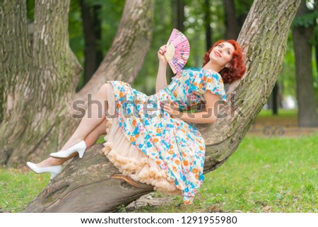charming girl with red hair in retro style posing in a vintage dress with a fan in her hands on a tree in the Park