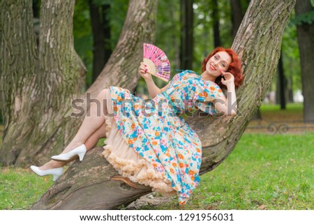 charming girl with red hair in retro style posing in a vintage dress with a fan in her hands on a tree in the Park