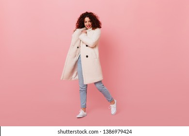Charming girl with red lipstick posing on pink background. Portrait of curly woman in white wool coat and light jeans