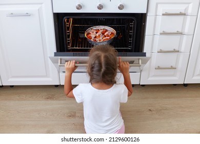 Charming girl sitting near gas stove on floor in kitchen and opening oven, curious female child waiting for cake or pie, baking in stove, homemade pastry.